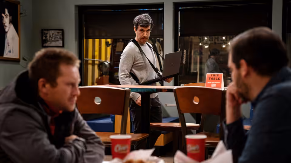Nathan Fielder with laptop approaches two men seated at a table