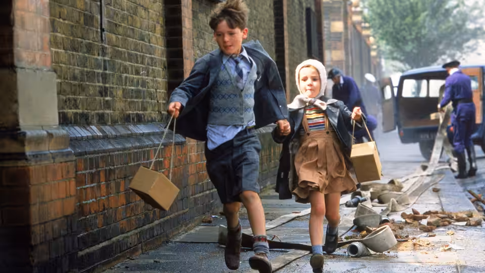 Bill (Sebastian Rice-Edwards) and Sue (Geraldine Muir) running down a rubble-strewn street carrying cases