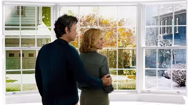 A man and woman stand embracing while looking out large windows at a snowy yard