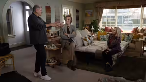 An older man stands gesturing toward another seated man while a woman watches in a living room