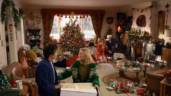 A woman holds a box while seated on the floor of a Christmas-decorated living room with family around