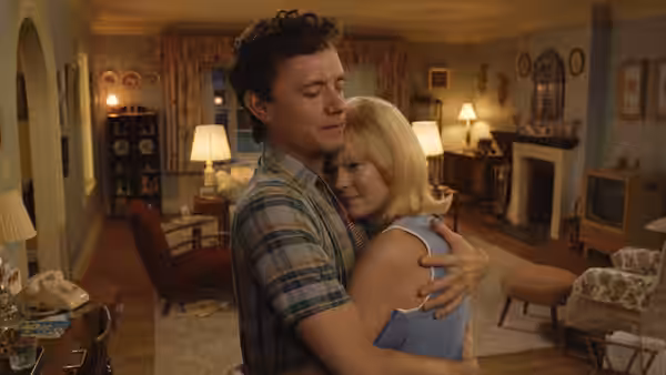 A man in a plaid shirt embraces a blonde woman in a living room