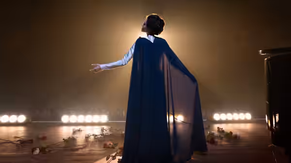 a woman in a blue cape stands with arms open on a rose-strewn stage