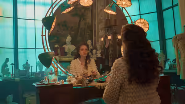 two women talk in an ornate dressing room filled with mirrors and lamps