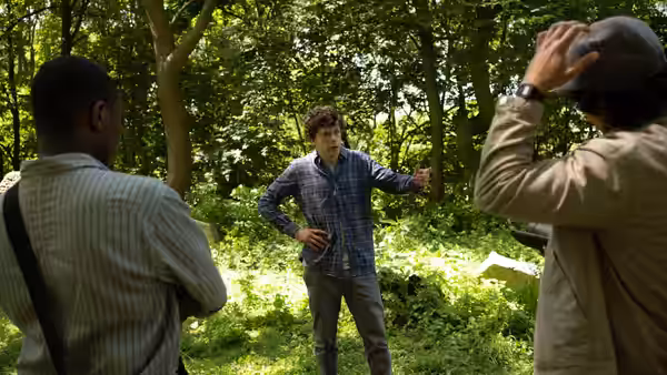 Man in plaid shirt gesturing while two others watch in woods