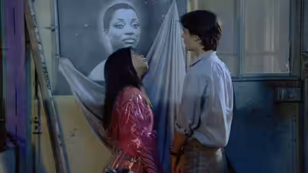 Alba and Jules stand before a large poster on a wall