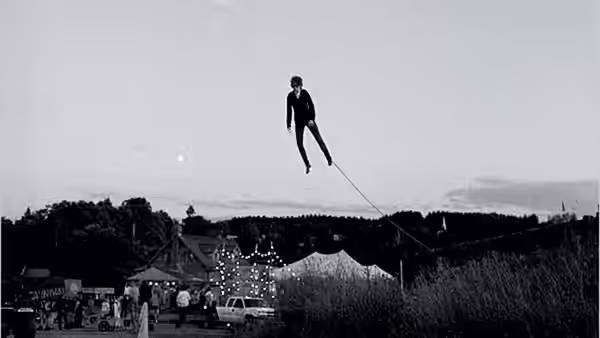 A lone figure hangs above a fairground, suspended from a taut line