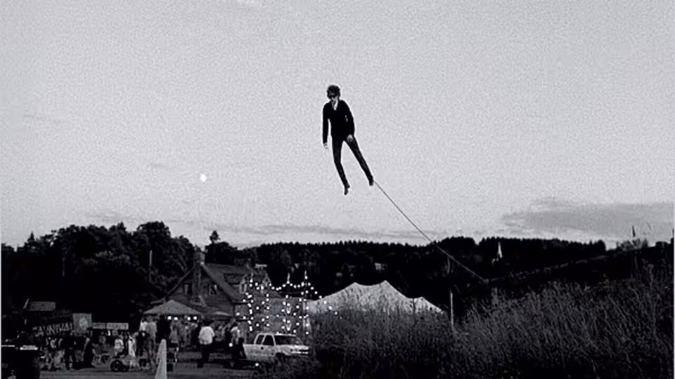 A lone figure hangs above a fairground, suspended from a taut line