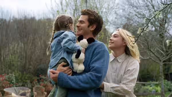 Tobias (Andrew Garfield) holds a young girl beside Almut (Florence Pugh) outdoors