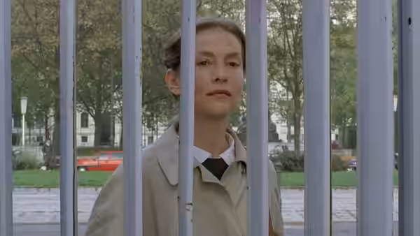 Erika Kohut (Isabelle Huppert) standing behind vertical metal bars outdoors