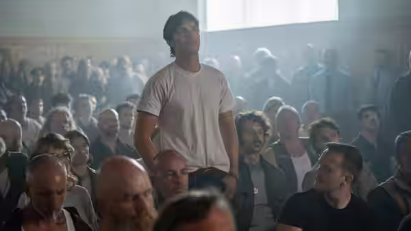 Robert Jay Mathews (Nicholas Hoult) standing among seated people in a hall