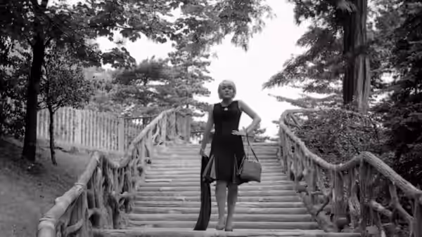 Cléo stands on a wooden staircase in a tree-lined park, holding a handbag