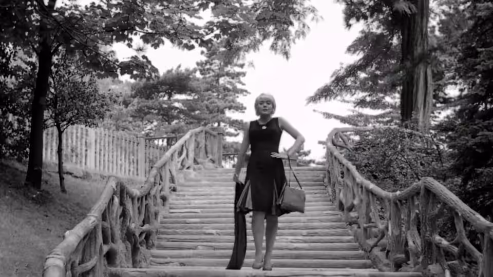 Cléo stands on a wooden staircase in a tree-lined park, holding a handbag