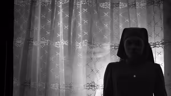 A nun stands before a patterned lace curtain in soft window light