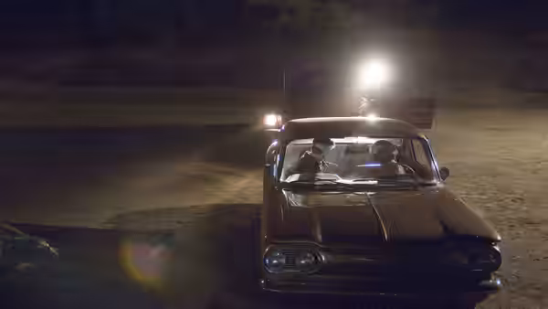 Two men sit inside a car at night with bright headlights on