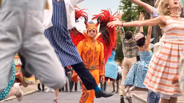 Performer in orange feathered costume dancing among a colorful street crowd
