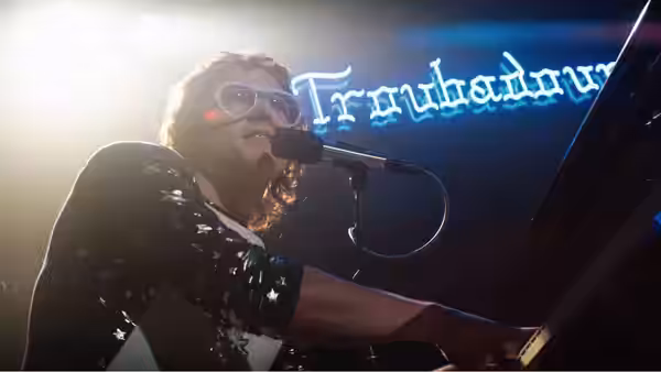 Elton John (Taron Egerton) singing at piano beneath Troubadour neon sign